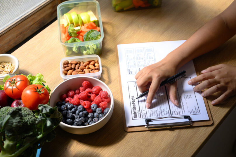 Nutritionist analyzing and writing a healthy meal plan with fresh fruits, vegetables, and nuts on the table.