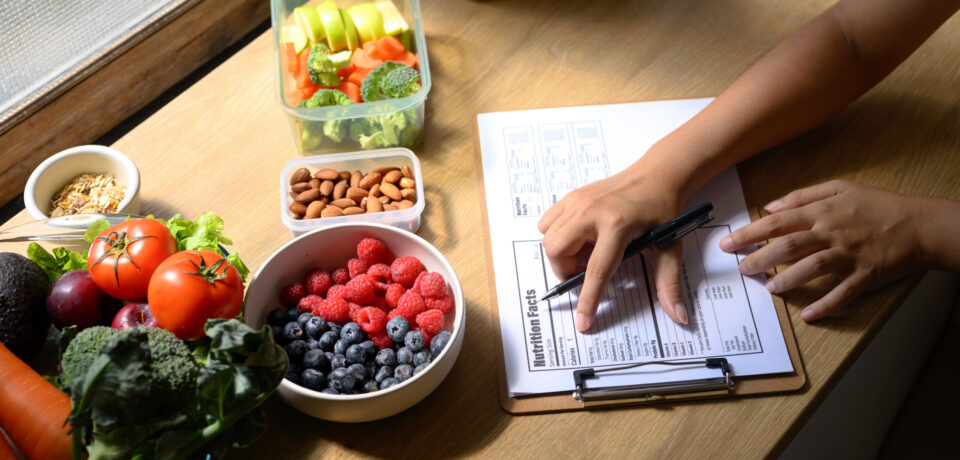 Nutritionist analyzing and writing a healthy meal plan with fresh fruits, vegetables, and nuts on the table Nutritionist analyzing and writing a healthy meal plan with fresh fruits, vegetables, and nuts on the table.